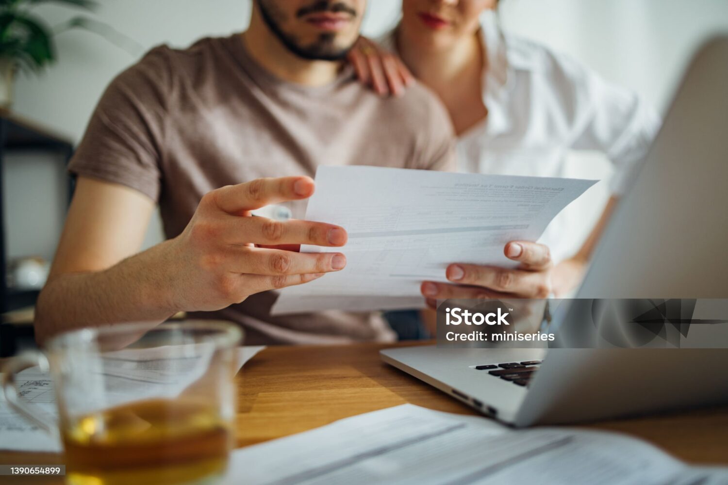 Close up photo of male hands holding bill and pen and female hands hugging him while they doing home finances together online on a laptop computer and drinking cup of tea in the kitchen.